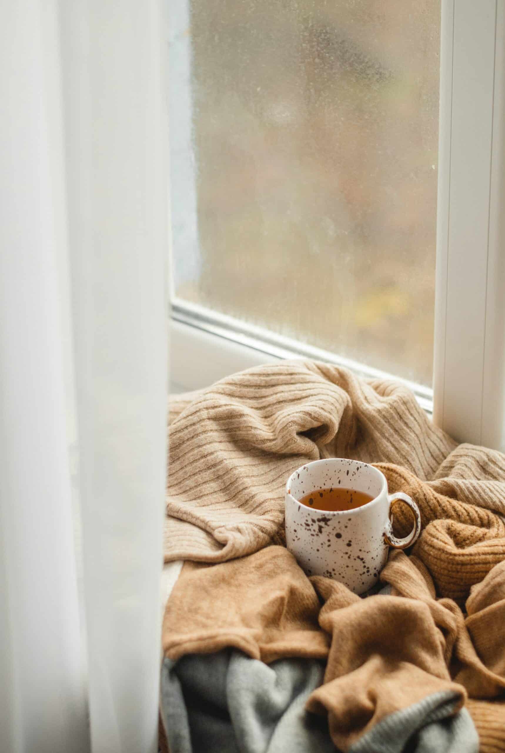 A ceramic mug of tea resting on soft brown knits beside a foggy window, creating a calming and cozy moment - perfect visual for slowing down and resetting ADHD brain fog.