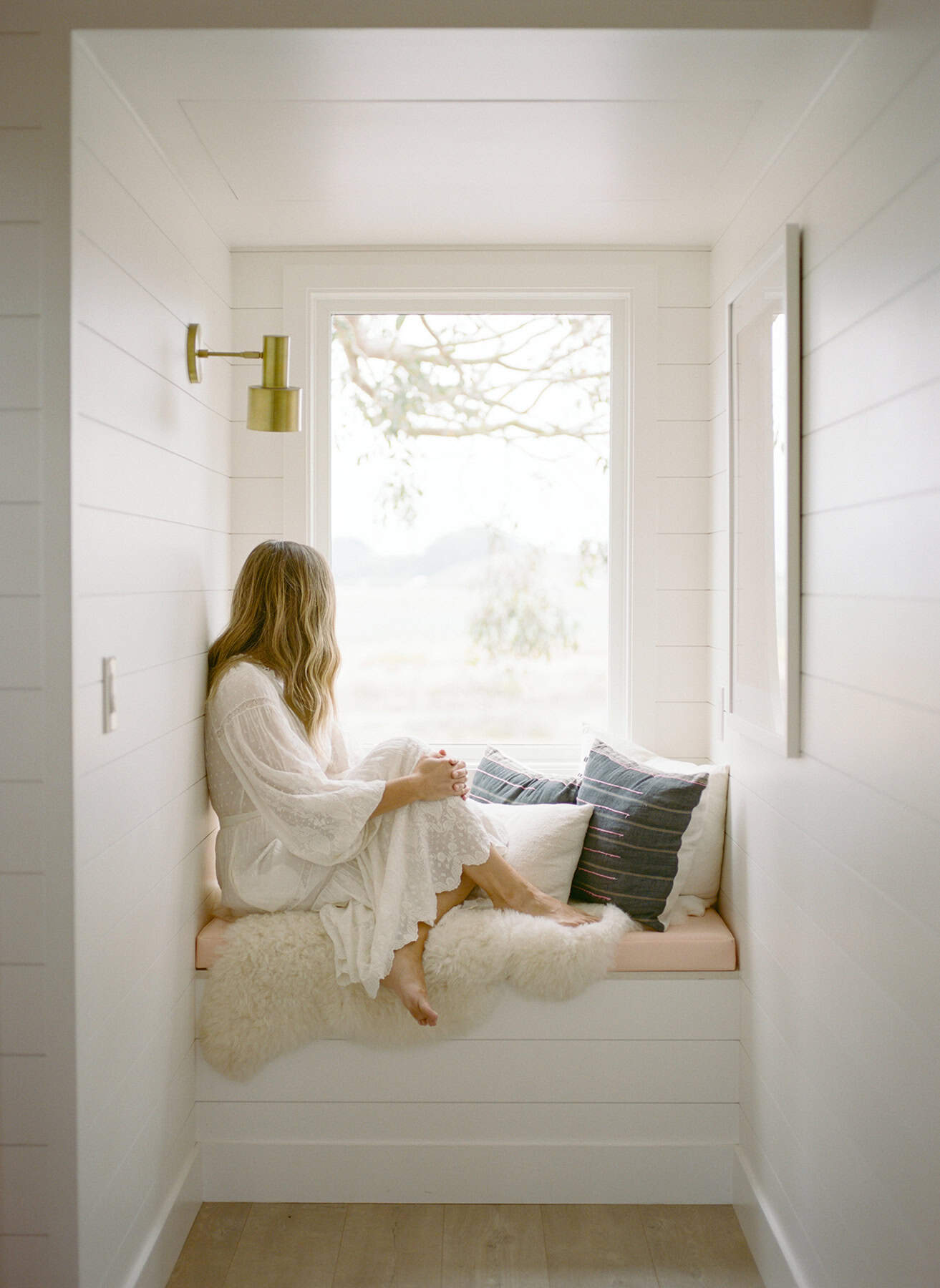 Woman in a white dress sitting quietly in a cozy window nook, looking outside on a soft, overcast day—capturing the stillness of learning how to function with depression.