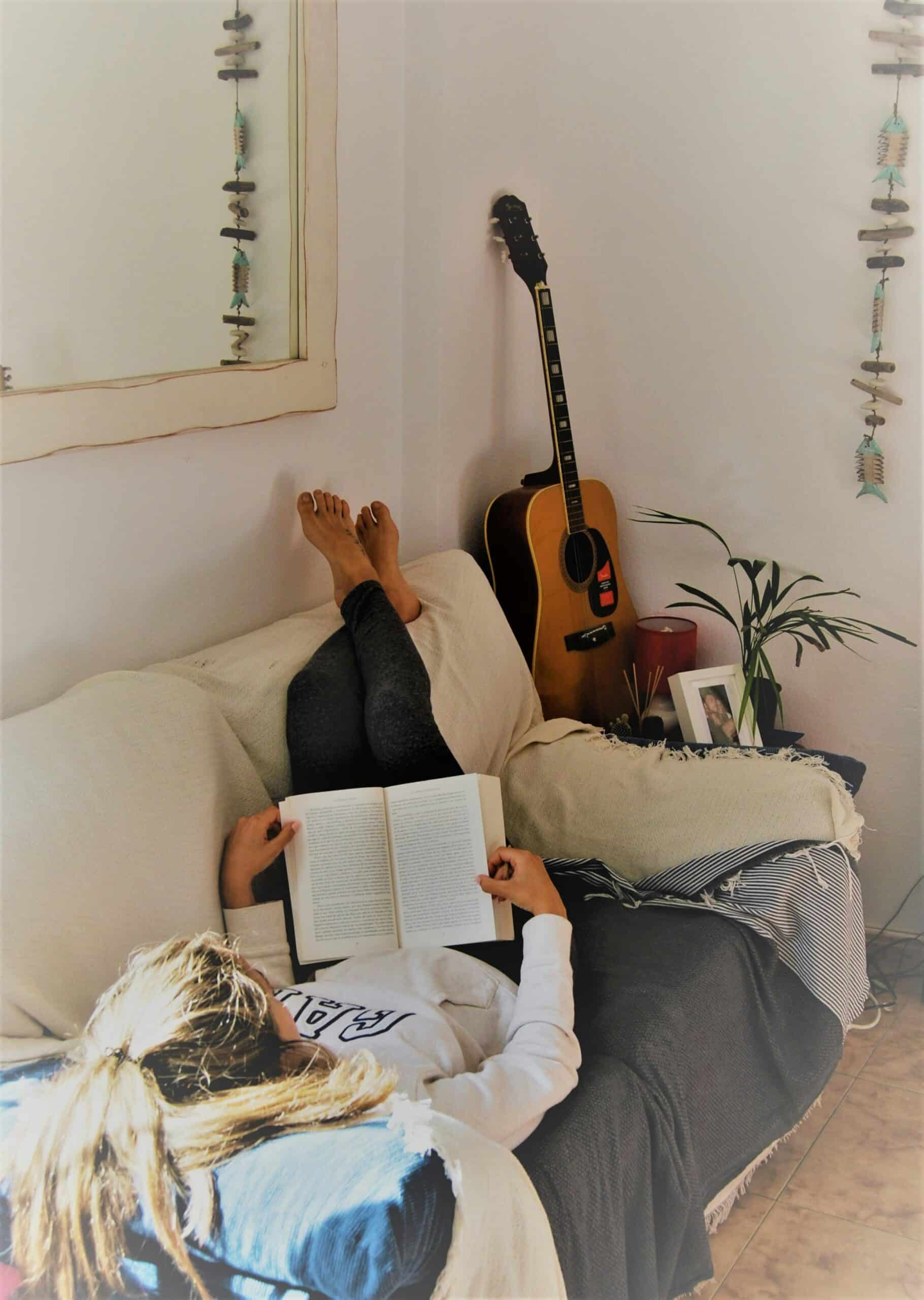 Woman reading a book on the couch, representing cozy self-care must-haves for relaxing at home.