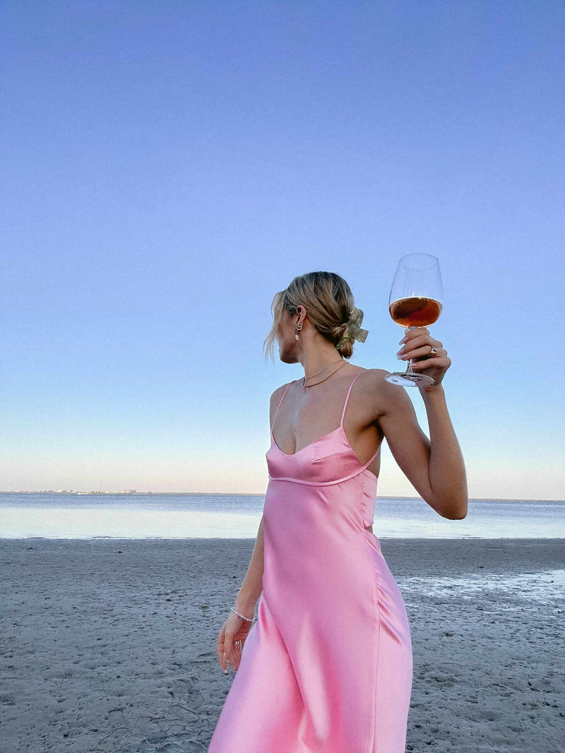 A woman in a pink dress holding a glass of wine on the beach, enjoying one of many fun ways to relax when stressed.