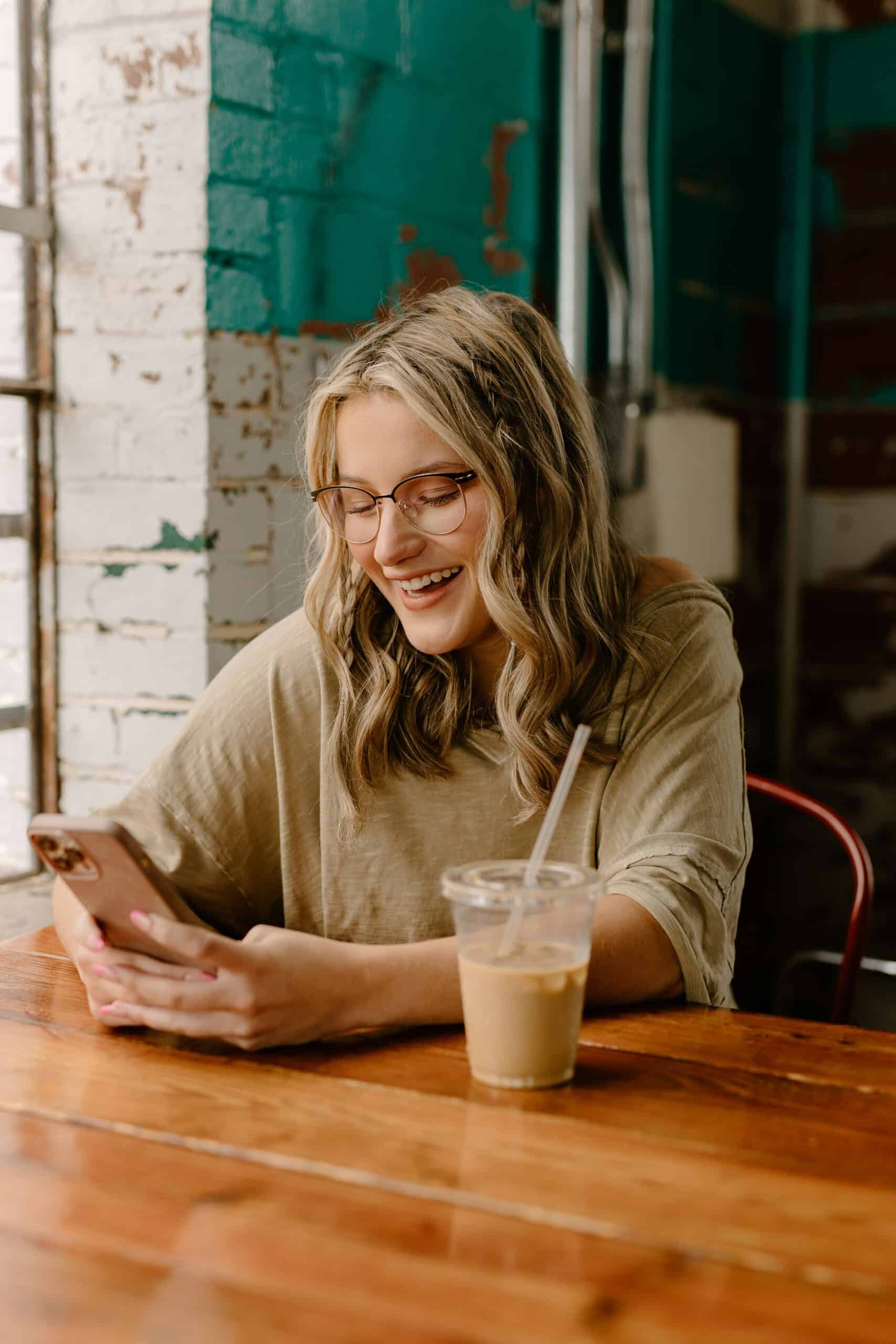 Young woman smiling at her phone in a coffee shop with an iced coffee, representing simple daily micro habits for productivity.