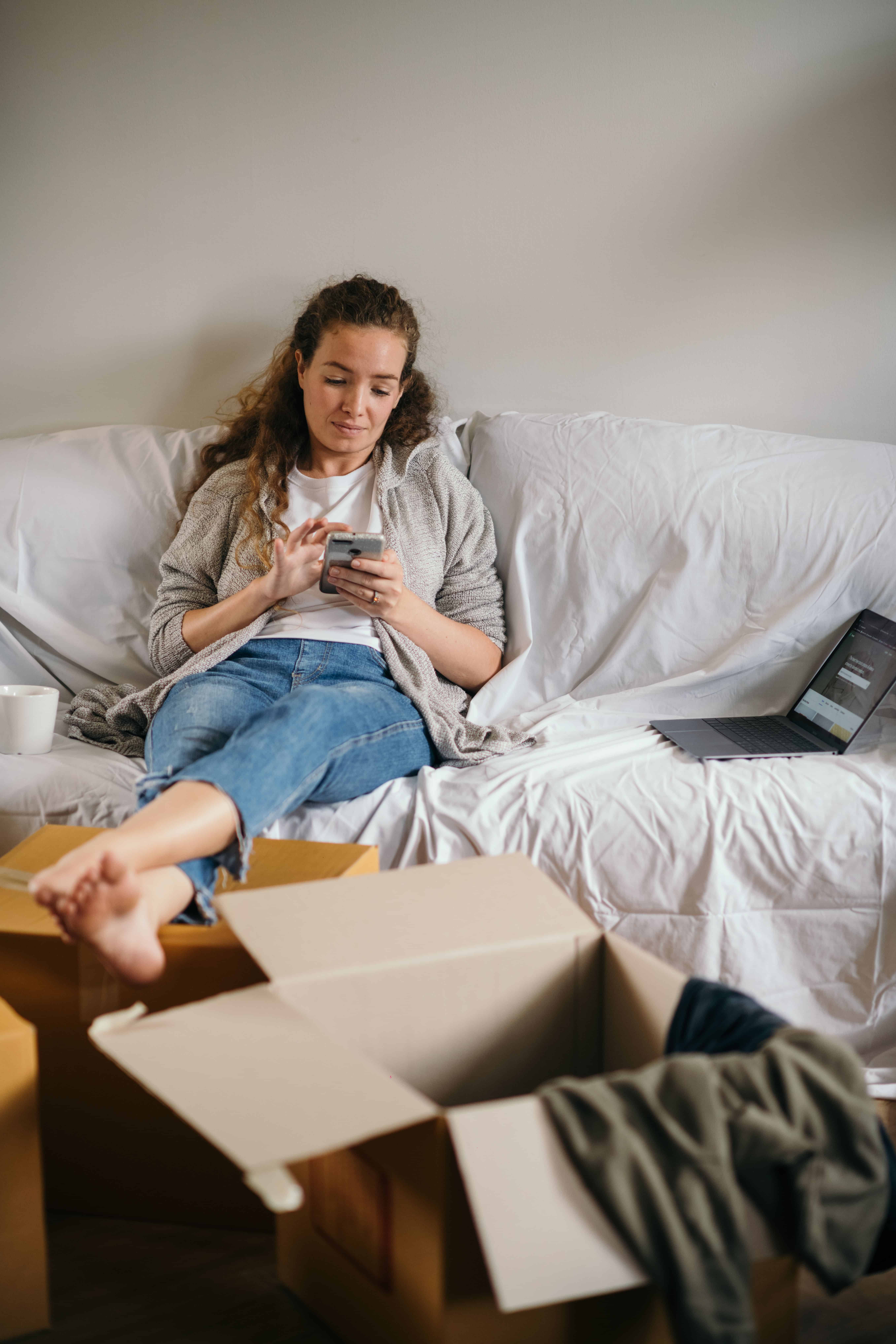 Woman with ADHD cleaning avoidance sitting on a couch using her phone beside boxes and household clutter